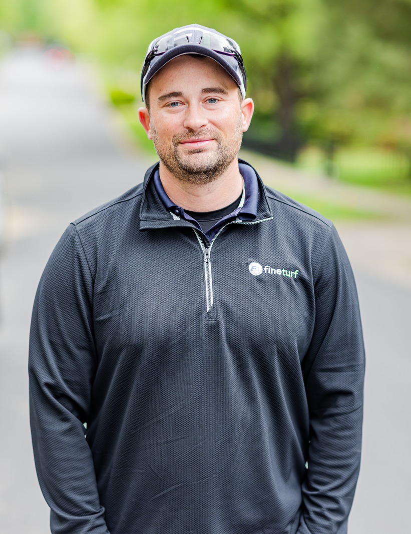 Branden Rheinschmidt, owner of FineTurf, smiling in a branded black polo shirt, standing outdoors in a residential area, embodying local expertise in lawn care.