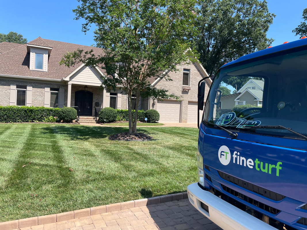 Lush green lawn with striped pattern, FineTurf lawn care truck parked in front of a residential home, showcasing professional lawn care services.